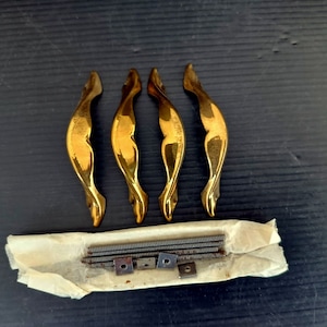 Puede incluir: Cuatro tiradores de puerta de armario de color dorado con un diseño curvo decorativo. Los tiradores se empaquetan con tornillos en un sobre de papel blanco.