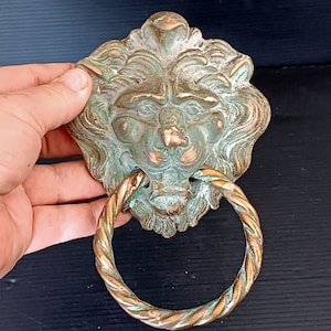 Op de afbeelding: Een bronzen deurklopper in de vorm van een leeuwenkop met een ringhandvat. Het gezicht van de leeuw heeft gedetailleerde kenmerken en een verweerde, verouderde uitstraling. De ringhandgreep is gedraaid en heeft een bronzen afwerking.