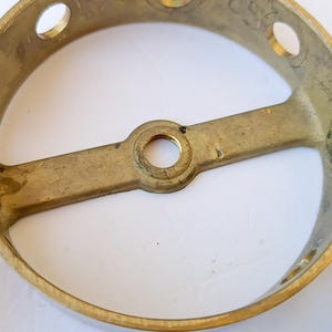 May include: A circular brass light fixture canopy with a metallic gold finish. The component features a central threaded hole on a horizontal crossbar and several evenly spaced circular openings along the outer rim for mounting purposes.