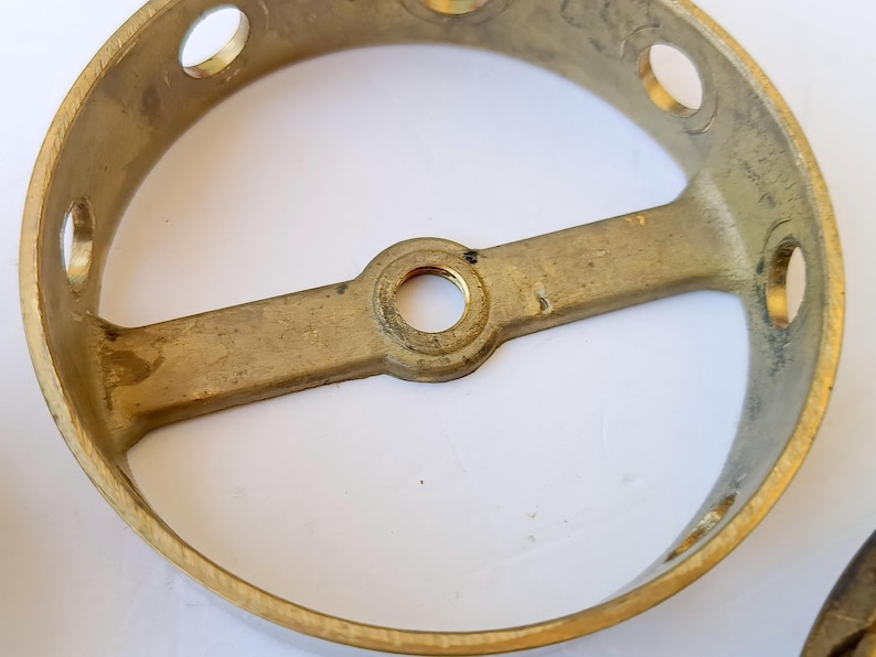 May include: A circular brass light fixture canopy with a metallic gold finish. The component features a central threaded hole on a horizontal crossbar and several evenly spaced circular openings along the outer rim for mounting purposes.