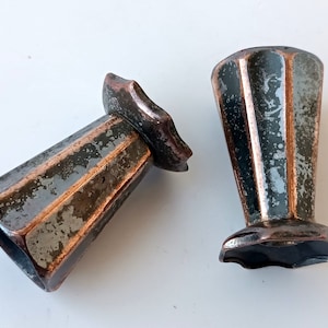 May include: Two antique metal vase feet with a dark, weathered finish and copper accents. The vase feet have a fluted design and a flared base. The metal has a distressed appearance, suggesting age and use. The vase feet are on a white surface.