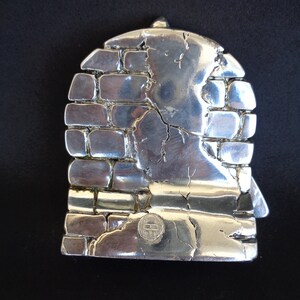 May include: Silver-toned metal sculpture of a brick wall with a textured surface. The sculpture has a circular shape and a small, circular stamp on the bottom.