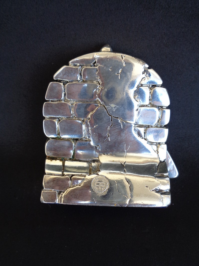 May include: Silver-toned metal sculpture of a brick wall with a textured surface. The sculpture has a circular shape and a small, circular stamp on the bottom.