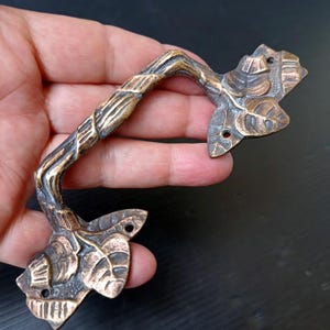 Peut inclure: Une poignée de meuble en bronze avec un design inspiré de la nature. La poignée présente une prise texturée en forme de vigne et des extrémités en forme de feuilles. La poignée mesure environ 15 cm de long.
