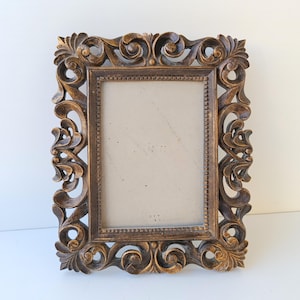 Op de afbeelding: Omlijste rechthoekige fotolijst met een gouden afwerking. Het frame heeft gedetailleerde krullen en bloemmotieven langs de randen. De binnenkant heeft een neutrale achterkant, klaar voor een foto of kunstwerk. Een decoratief woondecoratie-item.