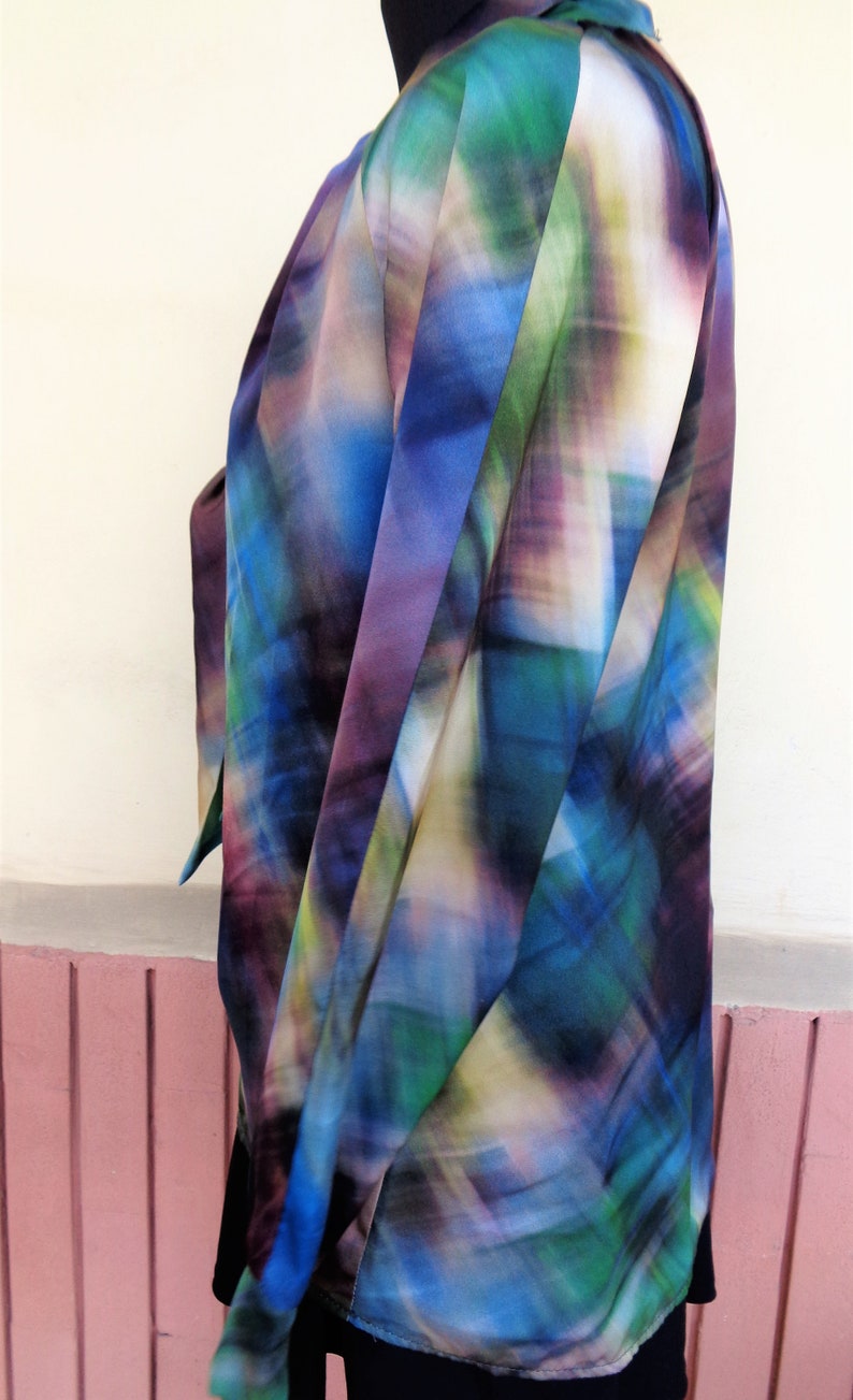 May include: A colorful, abstract-patterned blouse in shades of blue, green, purple, and yellow. The fabric drapes softly, suggesting a lightweight material. The blouse is worn over a black garment.