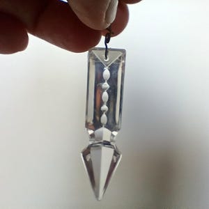 Peut inclure: Un prisme en cristal transparent avec une pointe pointue et une série de petites facettes rondes le long du centre. Le prisme est suspendu à un fil fin et noir.