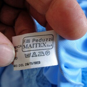 May include: White tag with black text that reads "Fil.li Peduzzi MAITEX S.p.A." and a 40 degree washing symbol, a triangle with an iron symbol inside, a square with a circle inside, and a circle with a P inside. The tag is attached to a light blue fabric.