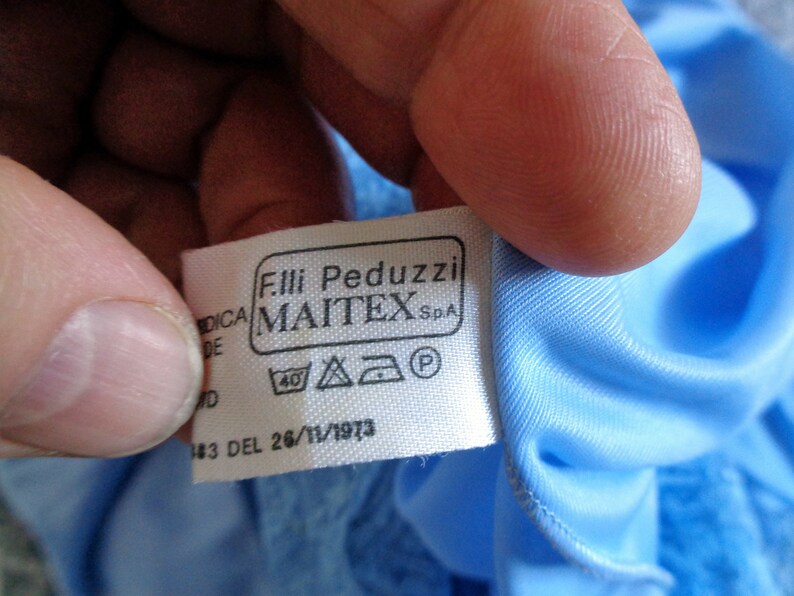 May include: White tag with black text that reads "Fil.li Peduzzi MAITEX S.p.A." and a 40 degree washing symbol, a triangle with an iron symbol inside, a square with a circle inside, and a circle with a P inside. The tag is attached to a light blue fabric.