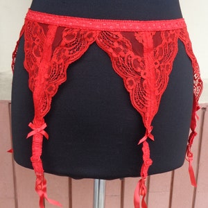 Può includere: Una giarrettiera in pizzo rosso con un design a bordo smerlato. La cintura presenta delicati dettagli in pizzo, spalline regolabili e piccoli fiocchi. La giarrettiera è esposta su un manichino nero.
