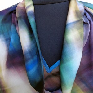 May include: A colorful blouse with a watercolor-like pattern in shades of blue, purple, and green. The blouse has a draped neckline and a flowing, lightweight fabric, perfect for a stylish and artistic look.