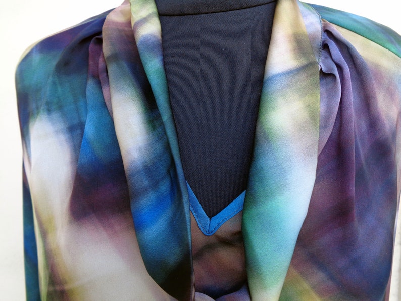 May include: A colorful blouse with a watercolor-like pattern in shades of blue, purple, and green. The blouse has a draped neckline and a flowing, lightweight fabric, perfect for a stylish and artistic look.