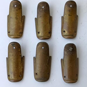 Op de afbeelding: Zes antieke messing sleutelgatdeksels. Elke afdekking heeft een rechthoekige vorm met een afgeronde bovenkant en twee zijvleugels. Ze hebben een klein gaatje aan de bovenkant en twee kleine gaatjes aan de onderkant. Het messing heeft een aangetaste, verouderde uitstraling.