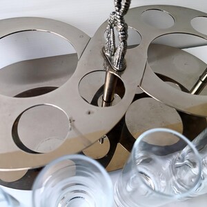 May include: A silver-toned metal bar caddy with circular cutouts for glasses. The caddy has a decorative handle and is designed to hold multiple glasses. Clear drinking glasses are visible in the foreground.