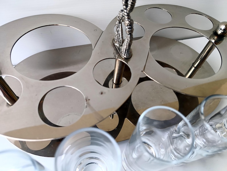 May include: A silver-toned metal bar caddy with circular cutouts for glasses. The caddy has a decorative handle and is designed to hold multiple glasses. Clear drinking glasses are visible in the foreground.