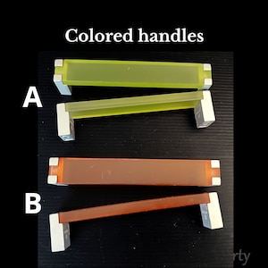 Op de afbeelding: Twee sets kastgrepen met gekleurde centra en zilverkleurige uiteinden. De bovenste set heeft een geelgroen centrum en de onderste set heeft een bruin-oranje centrum. De tekst "Colored handles" staat bovenaan.