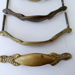 Puede incluir: Primer plano de cuatro tiradores de cajón antiguos de latón. Los tiradores tienen un diseño curvo con detalles decorativos en los extremos. Cada tirador tiene un tornillo roscado para el montaje. El latón tiene una pátina cálida y envejecida.