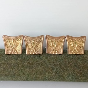 Könnte beinhalten: Vier goldfarbene Metall-Schubladenknöpfe in ausgestellter, trapezförmiger Form. Jeder Knopf weist ein erhabenes, geprägtes Blumen- und Blattmuster auf der Vorderseite auf, mit einer texturierten, punktierten Hintergrundoberfläche auf dem gesamten Beschlag.