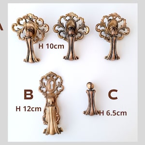 Könnte beinhalten: Verzierte, bronzefarbene Schrankbeschläge. Drei Teile sind 10 cm hoch, eines ist 12 cm hoch und eines ist 6,5 cm hoch. Die Beschläge weisen dekorative Schnörkel und ein Schlüssellochdesign auf einem Teil auf. Die Beschläge sind mit A, B und C gekennzeichnet.