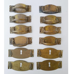 Op de afbeelding: Een verzameling antieke messing lade-grepen en sleutelgatplaten. De grepen hebben een gebogen handgreepontwerp met decoratieve hoekaccenten. De sleutelgatplaten hebben aan weerszijden van het sleutelgat een druppelvormig ontwerp.