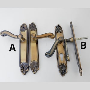 May include: Three antique brass door handles are displayed against a white background. Two handles are identical, featuring a long rectangular plate with ornate detailing and a curved lever. The third handle has a different design, with a keyhole and a different lever shape.
