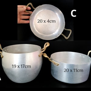 Puede incluir: Tres ollas de cocina de aluminio con asas de latón. Las ollas miden 20 x 4 cm, 20 x 11 cm y 19 x 17 cm. Se muestran sobre un fondo negro.