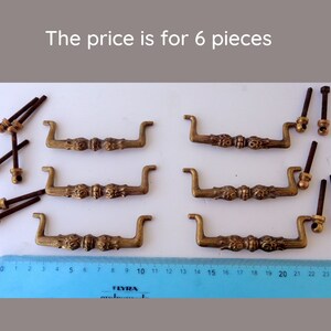 Op de afbeelding: Zes antieke messing lade handgrepen met decoratieve bloemmotieven. Elke handgreep heeft een gebogen handvat en wordt geleverd met montagemateriaal. Een liniaal is onder de handgrepen geplaatst. De tekst "The price is for 6 pieces" is zichtbaar.