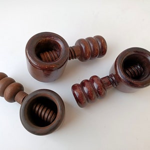 Op de afbeelding: Drie antieke houten notenkrakers met een donkerbruine afwerking. Elke notenkraker heeft een ronde kop met een schroefdraadmechanisme en een handvat met afgeronde groeven. De notenkrakers zijn gerangschikt op een witte ondergrond.