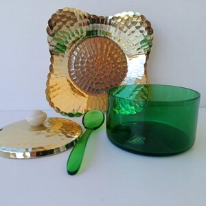 Könnte beinhalten: Ein dekoratives Servier-Set mit einem gehämmerten goldfarbenen Metallteller und Deckel, einer grünen Glasschale und einem kleinen grünen Löffel. Der Deckel hat einen weißen Knopf. Das Set ist zum Servieren bestimmt.