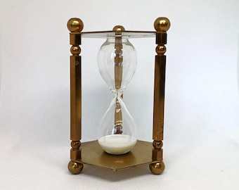 May include: An hourglass with a brass frame and clear glass bulbs. The hourglass is filled with white sand. The frame has a triangular base and is supported by four vertical posts with decorative ball accents.