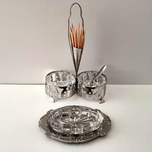Può includere: Un set da pranzo vintage con un portastuzzicadenti argentato con stuzzicadenti in legno, due piccole ciotole e un posacenere decorativo. Le ciotole hanno un design traforato e un cucchiaino. Il posacenere è in vetro trasparente.