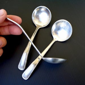 Peut inclure: Trois cuillères en argent avec des cuillères rondes et des manches décoratifs sont présentées sur un fond sombre. Une cuillère est tenue par une main. Les cuillères sont probablement destinées à servir ou à manger, et le design suggère un style vintage ou antique.