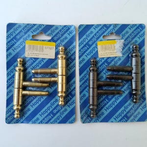 Puede incluir: Dos juegos de bisagras de puerta de metal empaquetados en tarjetas azules de marca. Un juego es de metal dorado pulido y el otro de metal color bronce oscuro. Cada bisagra presenta un cuerpo cilíndrico central con dos pernos roscados y remates.