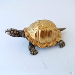 May include: A decorative turtle figurine with a gold-colored shell and silver-colored head, legs, and tail. The shell has a detailed, textured pattern. The figurine is a small, detailed sculpture.