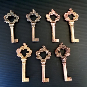 Op de afbeelding: Zeven sleutels in antieke stijl met een bronzen afwerking zijn gerangschikt op een donkere ondergrond. Elke sleutel heeft een decoratieve, opengewerkte kop en een lange steel. De sleutels variëren enigszins in kleur.