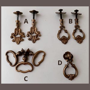 Könnte beinhalten: Eine Auswahl an antiken, bronzefarbenen Schrankbeschlägen. Das Bild zeigt vier verschiedene Stile: zwei Paare verzierter Griffe mit dekorativen Oberteilen, ein Satz von drei Griffen mit einem zentralen Griff und ein einzelner, eleganter Tropfgriff.