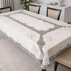 May include: A cream-colored rectangular tablecloth with a gray floral lace border and center design. The tablecloth is draped over a dark wooden table, with chairs visible around it. The room has a neutral color palette.