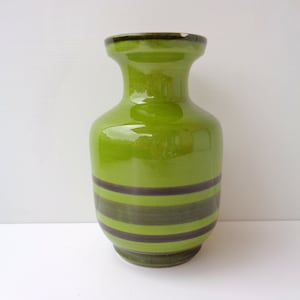 Könnte beinhalten: Eine grüne Keramikvase mit glänzendem Finish. Die Vase hat horizontale Bänder in Dunkelgrün und Braun. Sie hat einen breiten Sockel, einen abgerundeten Körper und einen ausgestellten Rand. Ein dekorativer Artikel.