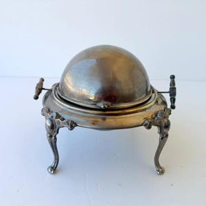 Puede incluir: Una fuente de servir antigua de color plateado con tapa abovedada y patas curvas ornamentadas. La fuente tiene dos asas de madera y un borde decorativo. El metal tiene un aspecto ligeramente deslustrado. Diseñada para mantener la comida caliente.