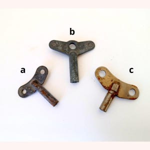 May include: Three vintage metal wind-up keys. Key 'a' is a dark grey with rust, key 'b' is a light grey with a butterfly-shaped head, and key 'c' is a light brown with rust. The keys are arranged on a white surface.