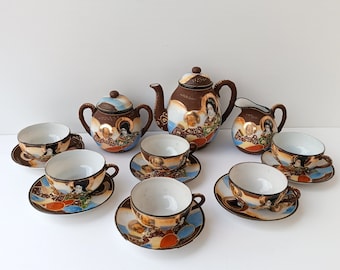 May include: A vintage Japanese tea set featuring a teapot, sugar bowl, creamer, and six teacups with saucers. The set has a brown background with hand-painted figures and gold accents. The teacups and saucers are approximately 4 inches in diameter.