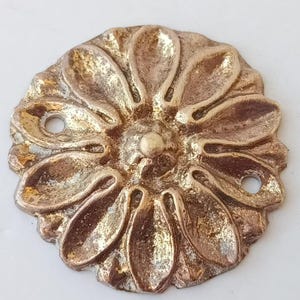 Könnte beinhalten: Ein kreisförmiges, goldfarbenes Zier-Medaillon mit einem erhabenen, zwölfblättrigen Blumendesign. In der Mitte befindet sich ein kleiner, abgerundeter Knopf, und an den gegenüberliegenden Seiten des Außenrandes befinden sich zwei kleine Löcher.