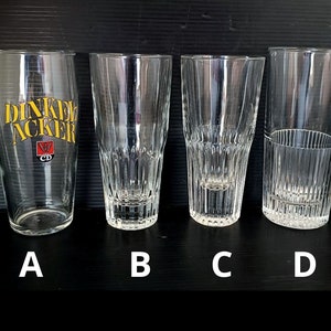 May include: Four clear glass drinking glasses. The glass on the left has the text "Dinkel Acker" printed on it. The glasses are arranged in a row from left to right, labeled A, B, C, and D.
