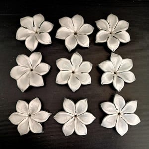 May include: Nine white, flower-shaped decorative objects with a glossy finish. Each flower has multiple petals and a dark center. The objects are arranged in a grid pattern on a dark surface.