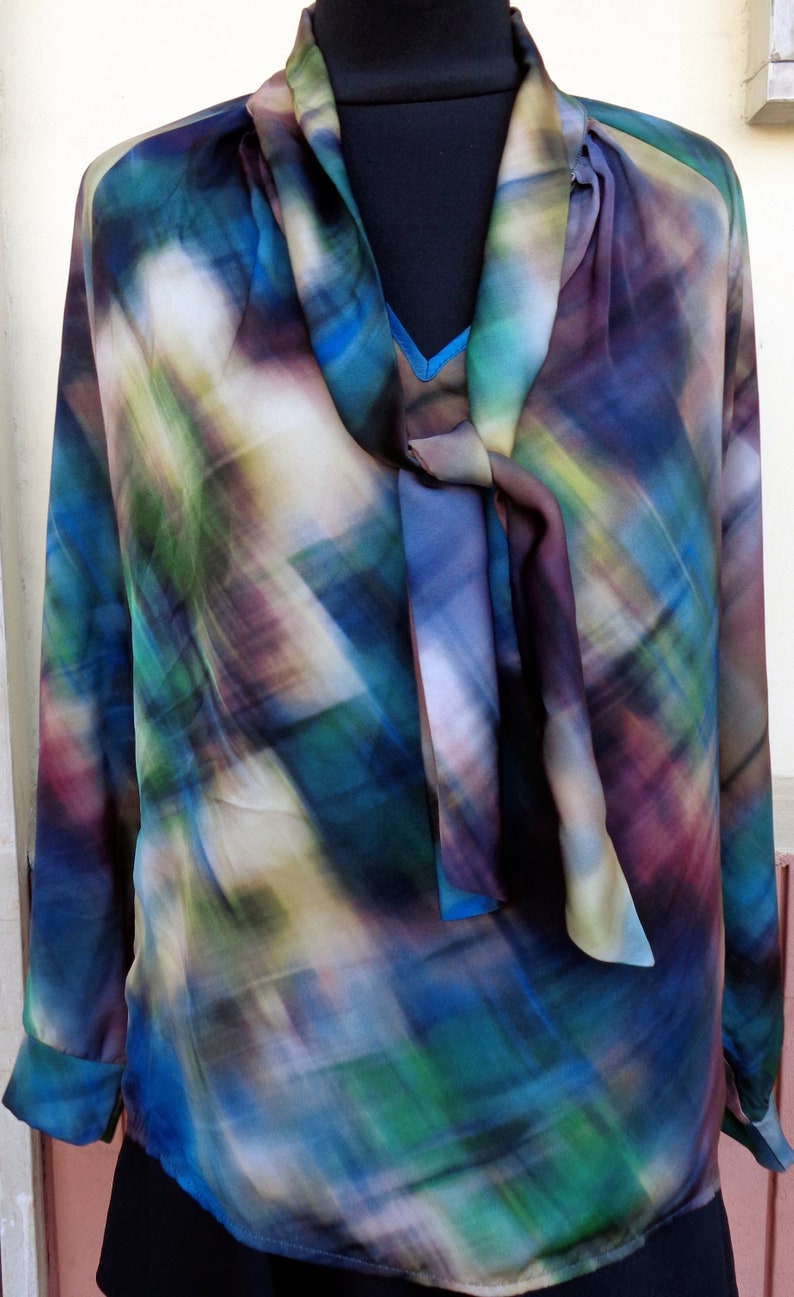 May include: A long-sleeved blouse with a colorful abstract pattern in shades of blue, green, purple, and yellow. The blouse features a tie-neck detail and is displayed on a black mannequin.