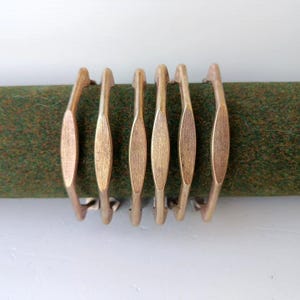 Può includere: Un set di sei maniglie per cassetti in metallo color ottone, ciascuna caratterizzata da un design sottile e allungato con un pannello centrale appiattito e estremità di montaggio curve, disposte in fila su una barra verde.