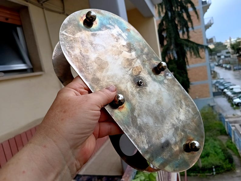 May include: A tarnished silver-colored serving tray with a handle and four rounded feet. The oval tray has a distressed finish with visible wear and tear. The handle is a curved, solid piece of metal. The tray is held by a person.