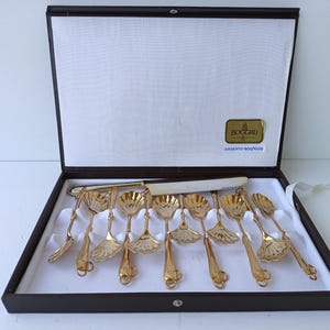 May include: A set of gold-toned metal spoons and a serving knife with shell-shaped bowls, presented in a dark brown rectangular case. The interior lid features a gold label with the text BOGGIALI and a blue label reading ARGENTO 800/1000.
