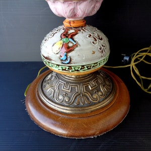 Op de afbeelding: Een keramische tafellamp met een roze, wit en groen bloemenontwerp op de bovenkant. De basis is rond, van hout met een gouden metalen rand. De lamp heeft een snoer en een stekker.
