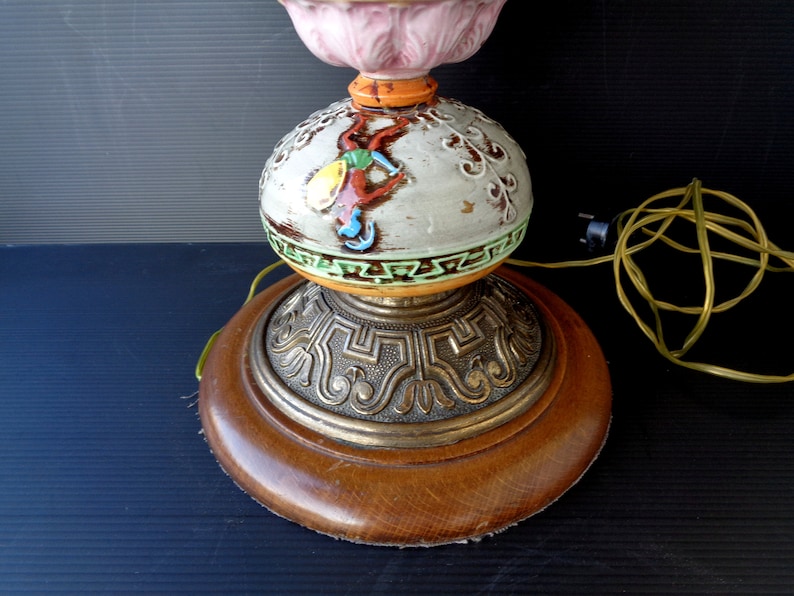 Op de afbeelding: Een keramische tafellamp met een roze, wit en groen bloemenontwerp op de bovenkant. De basis is rond, van hout met een gouden metalen rand. De lamp heeft een snoer en een stekker.